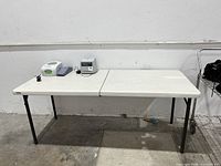 Front view of one folding table with items on top, showing white plastic textured top and black folding legs.