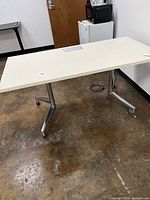Full view of cream rectangular training table desk on caster wheels with metal legs on brown floor near white walls.