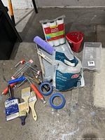 Photo showing the full set of painting tools including roller frames, covers, brushes, tape, trays, bucket, drop cloths, scrapers, and stir sticks laid out on floor