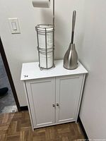 White vanity cabinet with double doors and small round knobs, showing brushed nickel toilet paper holder and toilet brush with caddy placed on top in a bathroom setting.