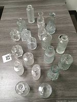 Top down view of 18 clear decorative glass vases and bottles on wood surface, showing varying shapes and textured patterns.