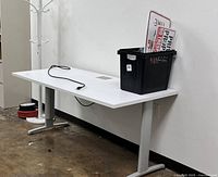 White rectangular office desk with silver metal legs and a built-in black power cord on the surface. A black plastic bin and some miscellaneous items are placed on the desk, which are not included.
