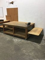 Front side view of wood rolling workbench with side shelf extended, two open storage compartments underneath, clamp on right side, caster wheels below.