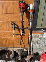 Photo shows three weed trimmers side-by-side outside against wood door and stone wall, red Craftsman, orange Husqvarna, green Yardworks.