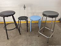 Four stools shown against a garage door on concrete floor: two tall stools with dark round padded seats and metal legs, one small black metal stool with holes in the seat, and one blue painted wooden stool.
