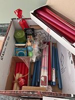 Box containing red taper candles, blue taper candles, pink taper candles, green pillar candle, party candles and small holiday shaped candles wrapped in plastic.