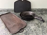Photo showing the large rectangular cast iron griddle, three cast iron fry pans stacked, and the heavy unknown purpose cast iron piece.