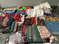 Wide view of a large assortment of Christmas quilting fabrics folded on a counter, showing a variety of prints and colors including green, red, blue, and cream backgrounds with festive holiday patterns.