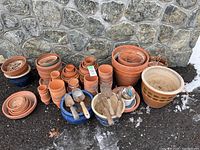 Wide view showing large group of terra cotta pots and trays arranged outside against stone wall along with blue glazed pots and gardening tools