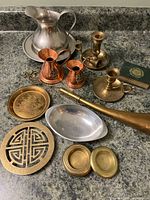 Photo of the lot's vintage brass, pewter, and copper items including large pewter pitcher, copper pitchers, candlesticks, trivet, horn and souvenir plate.