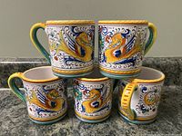 Five mugs arranged in two rows showing painted dragon motifs and colored rims