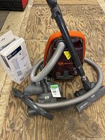 Full view of Sebo Airbelt K2 Volcano canister vacuum with hose, telescopic wand, primary floor tool, secondary floor tool, and opened box of replacement bags on wooden floor.
