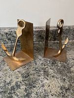 Pair of vintage brass anchor bookends shown side by side on countertop, showing front and side views for scale and detail.