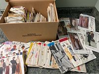 Many vintage sewing patterns in box and spread out on counter, showing variety of brands and styles with some Vogue Designer patterns visible