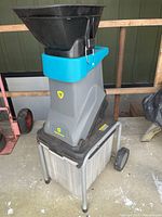 Front diagonal view showing large black hopper, grey body, blue accent, and attached plastic waste bin with wheels.