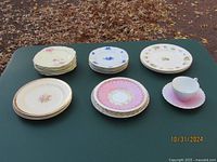 22 collectible plates and a cup & saucer arranged on a table, showing different floral and gold trimmed designs with varied colors including pink, blue and white.