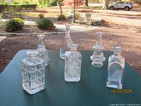 Six assorted glass or crystal decanters arranged on a green table outdoors showcasing their different shapes and etched patterns.