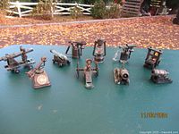 All 10 Play Me pencil sharpeners arranged in two rows on green surface outdoors with fall leaves in background. Mixed metal materials with rust and patina visible.