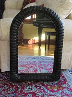 Full front view of arched mirror with black woven rattan frame standing on a patterned carpet with cream sofa behind it.