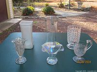 Overall photo showing five glass vases with different styles and finishes on outdoor table surface.