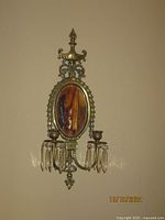 Front view of one brass wall sconce showing ornate design, oval beveled mirror, two candle holders with hanging crystal prisms