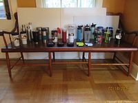 Wide view showing approximately 18 assorted water and thermos bottles arranged on a wooden bench, featuring various colors, sizes, and lid types for hot and cold beverages.