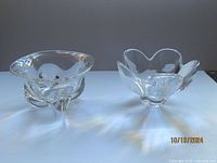 Two clear crystal bowls side by side, showing their shape and form. The left is the Edward Hald heavy art glass bowl with a rounded square base and flared edges. The right is the Lars Hellsten Pomona five-petal bowl with petal-like flared edges.