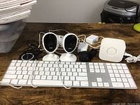 Photo showing two Apple wireless keyboards, two mini cameras on stands, Philips Hue Bridge with cables and power adapters on wooden table.