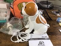 Beagle-shaped compressor nebulizer with attached tubing, mouthpiece, and parts in plastic bags, displayed with instruction manual and orange dog-graphic storage bag.