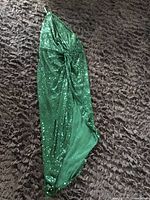 Photo showing green sequin gown laid flat on dark carpet with gathered center detail and one-strap design