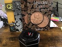 Three coin collection books with a black hexagonal tin container on a table, showing the front covers and the tin top.