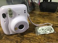 Front and slightly angled view of Fujifilm Instax Mini 11 camera in light purple with attached strap, also showing white earbuds coiled beside it.
