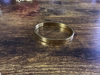 Gold tone rigid bangle bracelet laying flat on a wooden surface showing Roman numeral engravings on outer band.