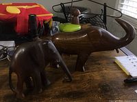 Two handmade wooden elephant carvings on a wooden surface. One is smaller and the other larger, with a leg missing that needs repair.
