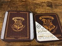 Photo showing brown box and tin case with Hogwarts crest and text indicating Harry Potter Hogwarts Playing Cards, suitable for ages 3+. Set new in box.