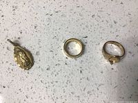Gold tone charm pendant and two rings placed on a white speckled surface; the charm has a loop for attaching; one ring features an oval design and foreign language markings inside band; the other ring is plain gold tone with no markings.