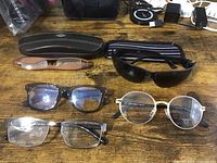 Five pairs of eyeglasses laid out on a wooden surface including reading glasses, sunglasses, and two hard eyeglass cases.