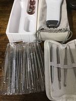 Photo of white facial scrubber device in a drawstring pouch with plastic tray holder beside it and plastic-wrapped metal skin extraction tools in fabric roll-up case.