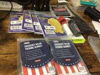 Photo showing two Wynne emergency mylar thermal blankets in sealed packaging with patriotic backing and multiple yellow emergency ponchos along with fire starter kit and flashlights.