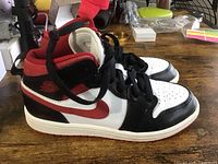 Side view of the Nike Air Jordan Mid 1 sneaker showing black, red, and white color scheme, leather, and laces
