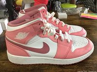 Side profile view of Nike Air Jordan 1 Mid sneakers in pink and white colorway on wooden surface, showing lateral side with Jordan wings logo on collar and Nike Swoosh.