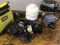 Overview of lot showing white Vornado circulator fan, gray bed pump device, and several electrical cords with adapters piled together on wooden table