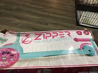 Box with partial image and product name 'Zipper Light-Up Scooter' with pink and blue colors and indication for kids 3+. The box corners are worn and creased.