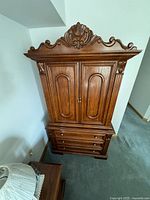 Full frontal view of the antique solid wood wardrobe showing intricate carved crown, two closed upper cabinet doors, and four drawers below with brass knobs.