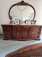 Front view of antique wooden vanity dresser with large oval mirror and multiple drawers and cabinet doors, with decorative items on top.