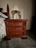 Front and side view of one wood bedside nightstand showing dark polished surface, three drawers with brass pulls, and carved decorative details.