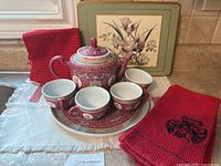 Full view of porcelain tea set including teapot, four cups with saucers, red woven table linens, and floral coasters
