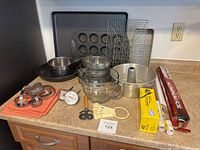 Full view of baking items arranged on kitchen counter including pans, bowls, racks, measuring spoons, cookie cutters, thermometer, and wrapped foil and paper rolls.