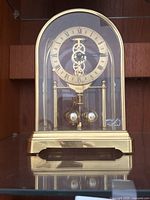 Front view of the gold colored Birks clock under a glass dome, showing Roman numeral ring and two pendulum balls at base.