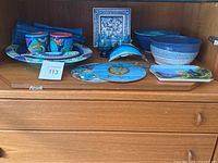 Photo shows variety of blue pottery plates, bowls, a small glass penguin figurine, a decorative square plate, and a rolled blue textile within a wooden shelf.